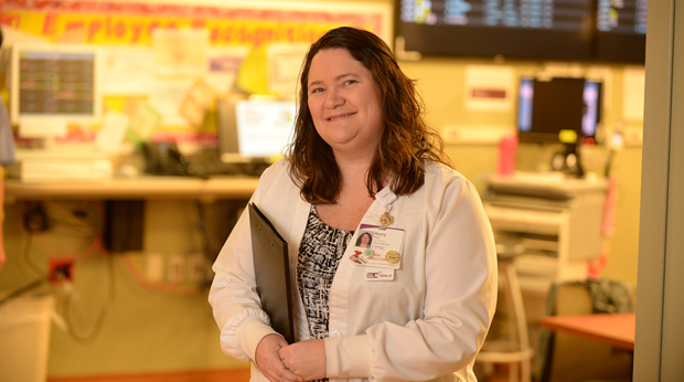 A nurse stands in a hospital hallway holding a clipboard.